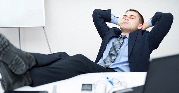 A man in a suit sitting at a desk, leaning back with his feet up and his arms behind his head