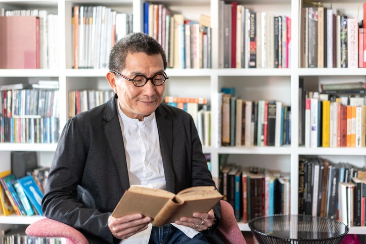 Photo of a man reading a book with bookshelves full of books behind him