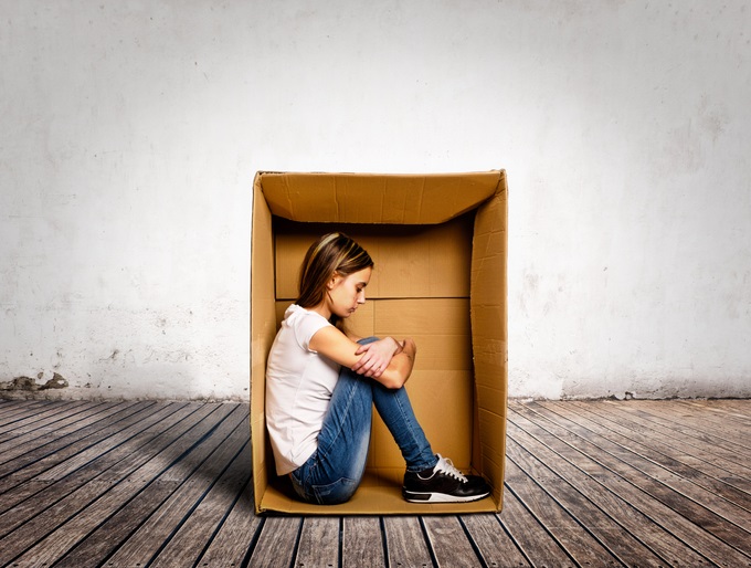 A woman sitting inside a cardboard box in the middle of the room. Her head is down and she looks sad and alone.