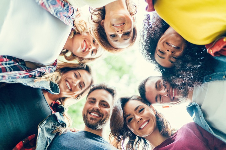 A group of friends standing in a circle with their heads together, looking down and smiling at the camera
