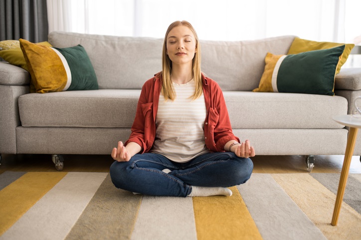A woman sitting on the floor with her legs crossed, eyes closed, and hands resting on her knees meditating