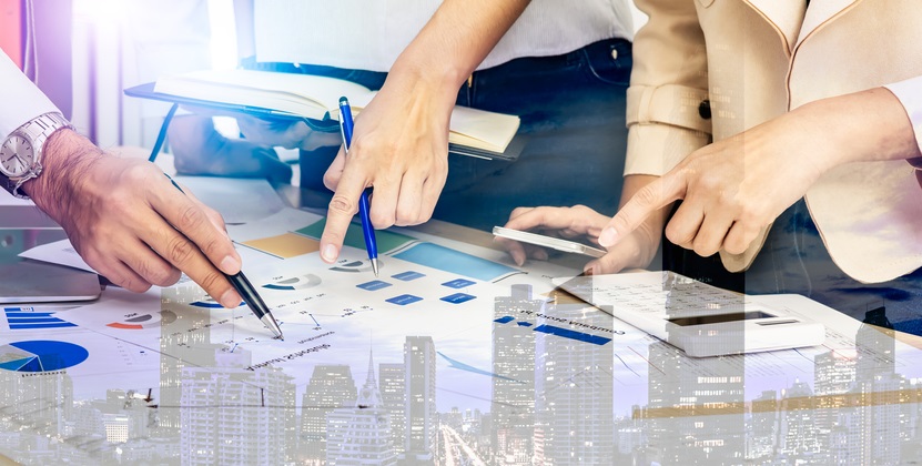 Photo of 3 people standing around a table pointing at charts and data