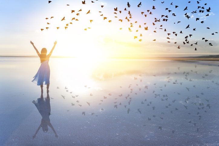 Photo of a happy woman with her hands up in the air. She is standing on the beach with the sun shining brightly and birds flying in the sky.