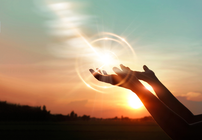 Two hands reaching up to the sky with a sunset in the background. There are light rays from the sun beaming around the hands.
